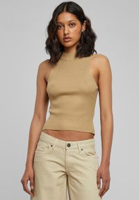 Ung kvinde med skulderlangt mørkt hår iført en ærmeløs beige ribbet top og lyse bukser, stående mod en ensfarvet baggrund.
