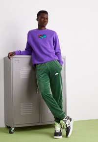 Lilla sweatshirt med et Nike-logo, parret med grønne velourbukser med et hvidt Nike-emblem og sorte sneakers med grønne accenter.