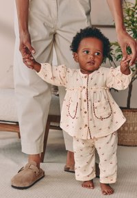 Tenue pour tout-petit de couleur crème en tissu léger, ornée de motifs de cerises rouges, avec des fermetures à boutons et deux poches plaquées.