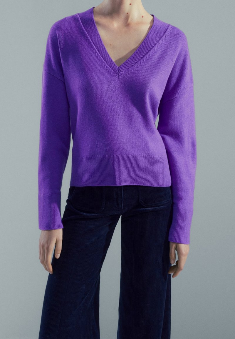 Personne portant un pull violet à col en V manches longues et un pantalon bleu foncé sur un fond gris uni.