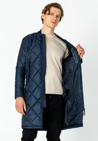 Marineblauer, gestepptes langes Jacket mit Reißverschluss, seitlichen Taschen und glattem Futter, getragen über einem hellbeigen Hemd und schwarzen Hosen.