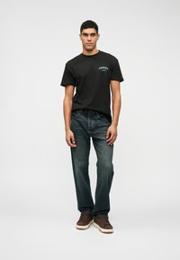 Juodas trumpomis rankovėmis T marškinėlis su mėlynu piešiniu ant krūtinės, derinamas su mėlynomis džinsinėmis kelnėmis ir rudais sportbačiais. Modelis stovi tiesiai prieš paprastą foną.