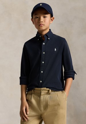 Mlad fant v mornarsko modri srajci z gumbi in zavihanimi rokavi, mornarskem pokrivalu in bež hlačah stoji ob enobarvnem ozadju.