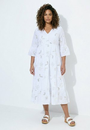 Frau mit lockigem Haar, die ein weißes, langärmliges, knöchellanges Kleid mit floralen Cutout-Mustern und weißen Plateausandalen trägt.
