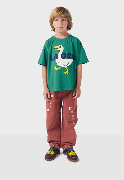 Zelené bavlnené tričko s bielym grafickým zobrazením kačky a modrým nápisom, v kombinácii s prehnane veľkými hnedo-červenými nohavicami so bielymi textovými akcentmi.