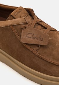 Brown suede shoe with a rounded toe, lace-up design, and a branded tag. Features a beige rubber sole and contrasting stitching details.