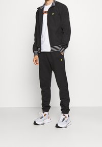 Zwarte zip-up jack met witte gestreepte manchetten, gecombineerd met bijpassende zwarte joggers. Heeft een gele logo op beide stukken en witte sneakers.