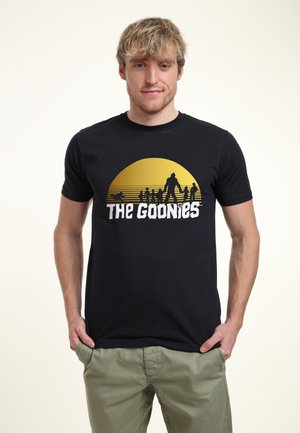 Lyshåret mand iført sort T-shirt med gul sol og sorte silhuetter af mennesker, tekst "THE GOONIES" foran, kombineret med lysegrønne bukser.