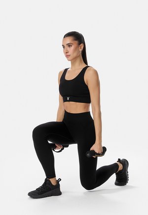 Naine mustades treeningriietes sooritab sammudes väljaasteid, hoides käes väikseid hantleid, taustal valge tasane sein.