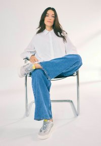Chemise blanche, jean large bleu, et baskets grises avec des accents jaunes, assis sur une chaise en chrome contre un fond clair.