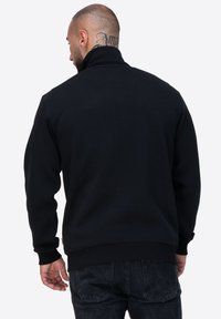 Felpa nera in pile con colletto alto, maniche lunghe e polsini e orlo a coste. Presenta un'etichetta logo discreta sulla vita.