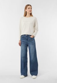 Krémová košile s dlouhými rukávy a nápisem "MINIMALIST", spárovaná s volnými modrými džíny. Oblečení doplňuje ležérní obuv. Čistý, jednoduchý design.