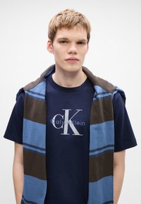 Jeune homme aux cheveux courts et clairs portant un t-shirt Calvin Klein bleu marine et un pull rayé bleu et brun drapé sur ses épaules.