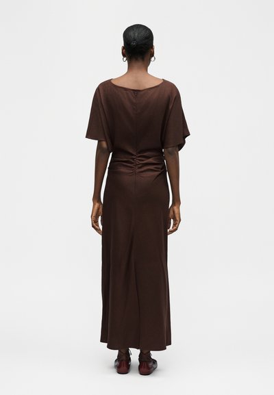 Γυναίκα με σκούρα μαλλιά μαζεμένα σε κότσο, φορώντας ένα μακρύ καφέ φόρεμα με κοντά φαρδιά μανίκια, στέκεται με το πρόσωπο στραμμένο προς τα πίσω σε ένα απλό φόντο.