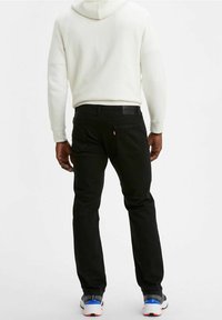 Jeans de mezclilla negros con un ajuste slim, bolsillos traseros visibles y un pequeño logo rojo. Combinados con una sudadera con capucha blanca y zapatillas deportivas.