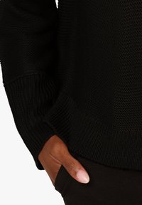 Černý svetr s texturou, ribovaný na rukávech a lemu, s volným střihem. Ruka položená v kapse černých kalhot.