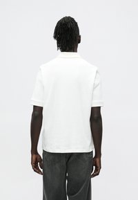 Osoba s tmavou pokožkou a dredy, oblečená v bílé polokošili s krátkým rukávem a vybledlých černých džínách, stojící před světlým jednobarevným pozadím.