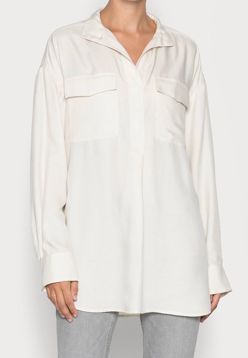 Camisa de manga larga de color beige claro con cuello, dos bolsillos en el pecho y un corte relajado. Fabricada con un tejido suave con un sutil brillo y con aberturas laterales.