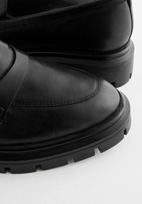 Mocassins en cuir noir avec semelles en caoutchouc épaisses, coutures détaillées et finition lisse et polie sur fond blanc.