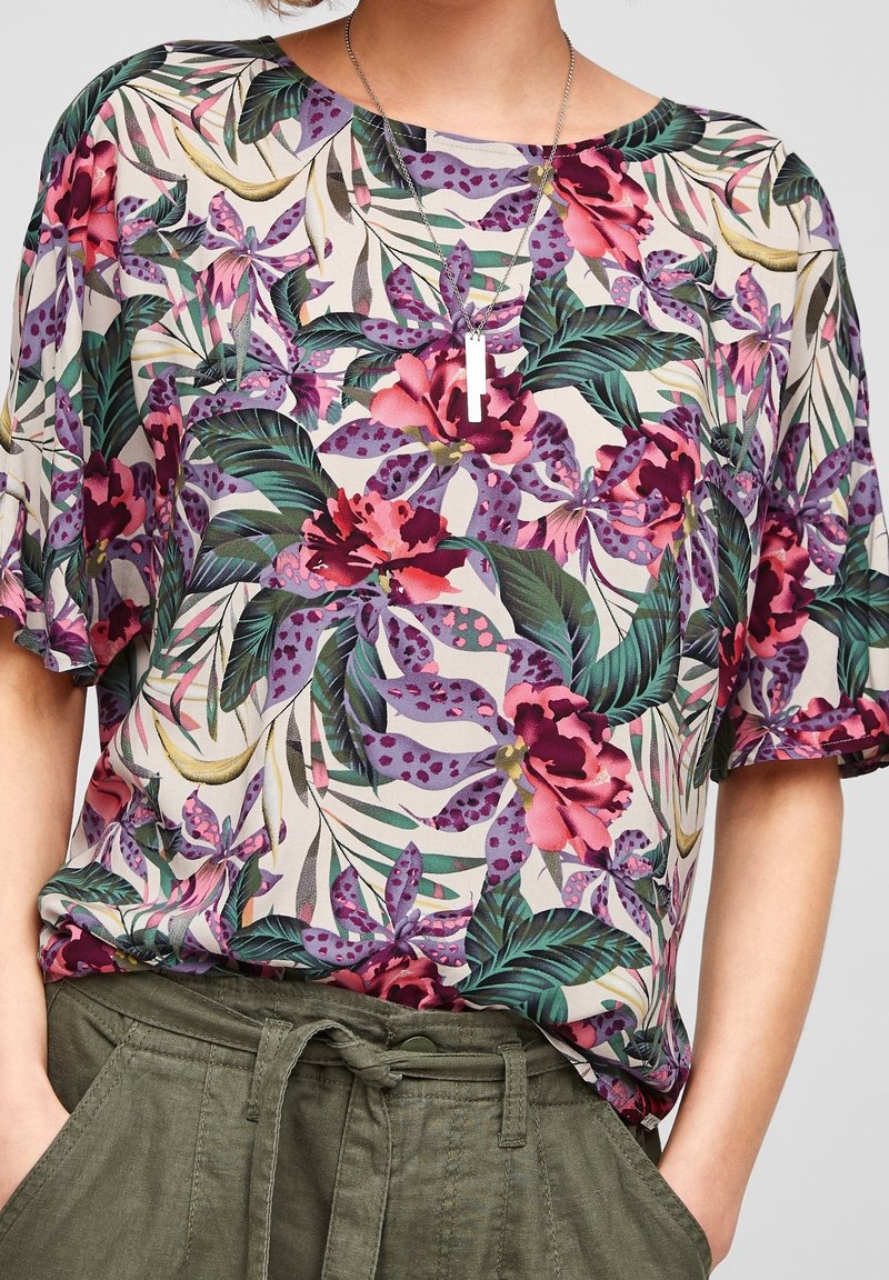 Top con stampa floreale con foglie viola, rosa e verdi su sfondo beige. Tessuto leggero con maniche corte e vestibilità ampia.