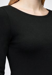 Gros plan d’une personne portant un haut noir à manches longues avec un col large et rond sur un fond neutre.