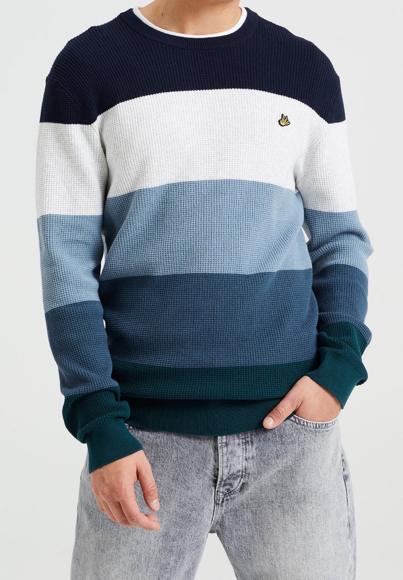 Hombre vistiendo un suéter de punto de manga larga con rayas horizontales en marino, blanco, azul claro, azul oscuro y verde azulado, combinado con jeans de color gris claro.