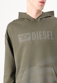 Olívovo zelená mikina s kapucňou vyrobená z mäkkej tkaniny, s predným klokanním vreckom a výrazným sivým logom "DIESEL" na hrudi.