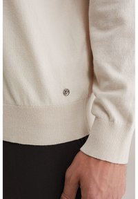 Pull de couleur crème en tissu tricoté doux avec un ourlet et des poignets côtelés. Présente un petit emblème près du bas à gauche.