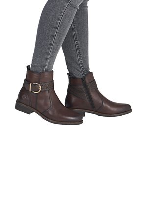 Braune Leder-Ankle-Boots mit einem dekorativen Riemen und einer goldenen Schnalle, mit einer glatten Oberfläche und einem seitlichen Reißverschluss, kombiniert mit grauen Jeans.