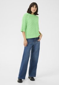 Lichtgroene, gebreide trui met korte mouwen en een ruitpatroon, gecombineerd met wijde blauwe denim jeans en zwarte schoenen.