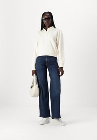 Maglione beige chiaro con collare, abbinato a jeans a gamba larga blu scuro. Il modello tiene una borsa beige, indossa occhiali da sole neri e sneaker bianche.