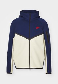 Marineblå og cremefarvet zip-up hoodie med høj krave, med sorte detaljer og et rødt Nike-logo på venstre bryst. Inkluderer en sidelomme.