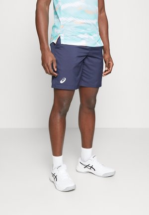 Homme portant un short de sport bleu marine, une chemise bleu clair à motifs, des chaussettes blanches Asics et des chaussures de course blanches Asics, debout sur un sol blanc.