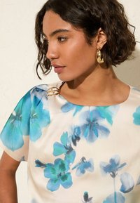 Blusa floral de satén suave con grandes flores azules y verde azulado sobre un fondo crema. Presenta un escote redondeado y un acento dorado en el hombro.
