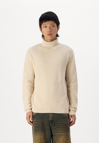 Beige gestrickter Rollkragenpullover mit einem strukturierten Muster. Gerippte Bündchen und Saum, lockere Passform, kombiniert mit dunklen, weit geschnittenen Jeanshosen.