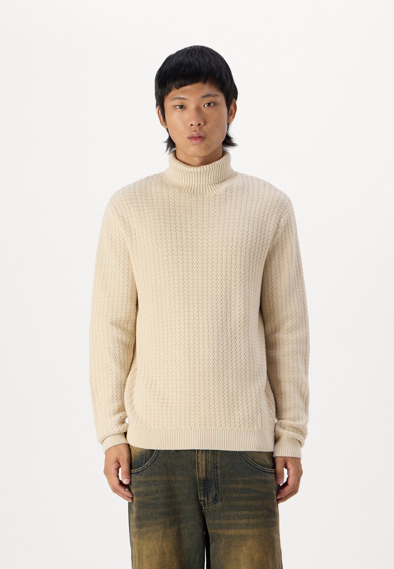 Beige gestrickter Rollkragenpullover mit einem strukturierten Muster. Gerippte Bündchen und Saum, lockere Passform, kombiniert mit dunklen, weit geschnittenen Jeanshosen.