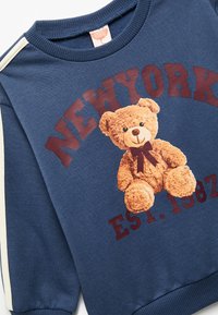 Navyblauwe sweater met een teddybeerafbeelding met een strik, verfraaid met bordeauxrode tekst "NEW YORK EST. 1982" en contrasterende crèmekleurige strepen.