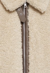 Giacca in pile beige con una texture morbida, caratterizzata da un'apertura con zip in pelle marrone e un colletto classico. La zip è di colore argentato.