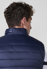 Chaleco de plumas azul marino con paneles acolchados horizontales, cuello alto con logotipo en relieve y una textura lisa y brillante.
