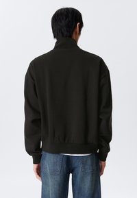 Une personne avec des cheveux noirs courts porte un sweat à col montant noir et un jean bleu, se tenant de dos contre un fond uni.