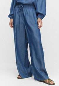 Denim vida byxor med elastisk midja och dragsko, med en slät textur och en djup blå färg, kombinerade med platta sandaler.