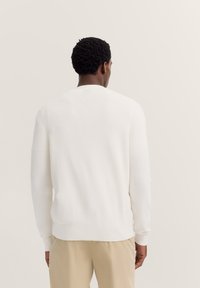 Weißer Strickpullover mit langen Ärmeln, Rundhalsausschnitt, geripptem Textil und enganliegenden Bündchen, getragen über beigen Hosen. Von hinten gesehen.