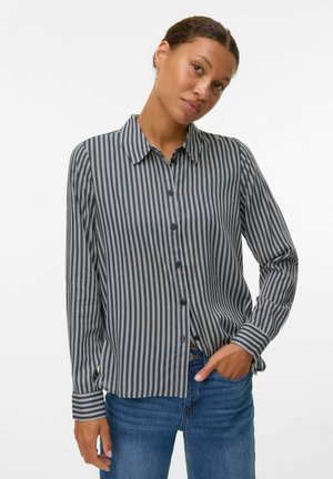 Donna con i capelli raccolti che indossa una camicia a righe verticali bianca e nera e jeans blu, posando con una mano in tasca.