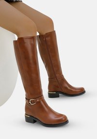 Botas de piel marrón hasta la rodilla con una textura suave, cremallera lateral y una hebilla decorativa en el tobillo. Tacón grueso para mayor estabilidad.