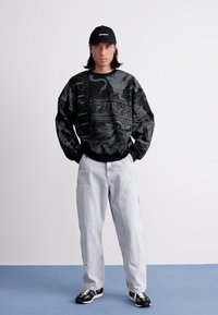 Young person wearing black graphic sweatshirt, white pants, black cap, and black-and-white sneakers standing against white background.