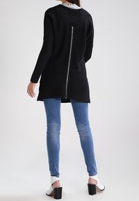 Frau, die einen schwarzen gestrickten langen Cardigan mit einem Reißverschluss auf dem Rücken, blaue Skinny-Jeans und weiße offene Blockabsatzschuhe trägt, vor einem einfarbigen Hintergrund steht.