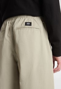 Pantalon beige avec une taille élastique, comprenant une poche arrière et une petite étiquette de logo noire à l'arrière. Tissu lisse et léger.