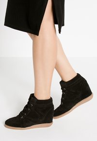 Jambes portant des baskets compensées noires avec lacets et semelles beige, associées à une jupe noire avec une fente à l’avant.