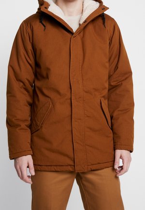 Personne portant une veste d’hiver marron à capuche avec une doublure polaire blanche et des poches avant, associée à un pantalon marron assorti.