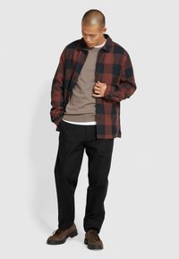 Camicia a scacchi rossa e nera, abbinata a un maglione marrone sopra una camicia bianca, pantaloni neri e scarpe marroni. Look casual a strati.